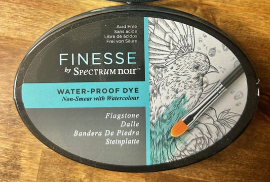 A round, dark grey Crafter's Companion Spectrum Noir Finesse water-proof dye ink pad in the color "Flagstone". The ink pad is acid-free and designed for use with watercolour media as it is water-proof.