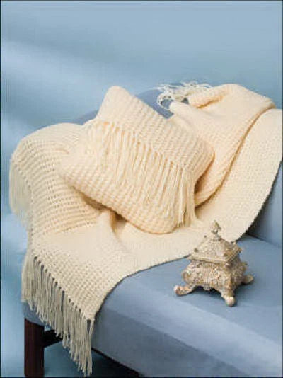 Cream-colored knitted blanket with fringe draped over a chair against a light blue background