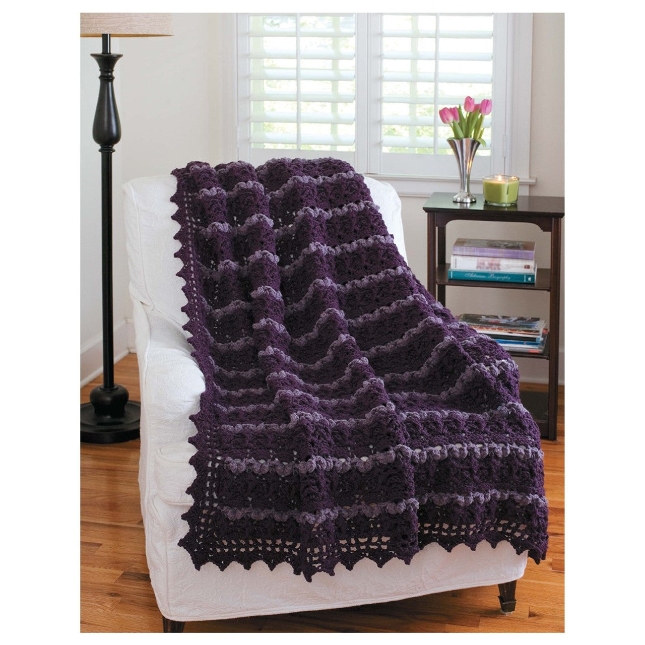 Purple crocheted blanket draped over a white chair in a room with a lamp and window.