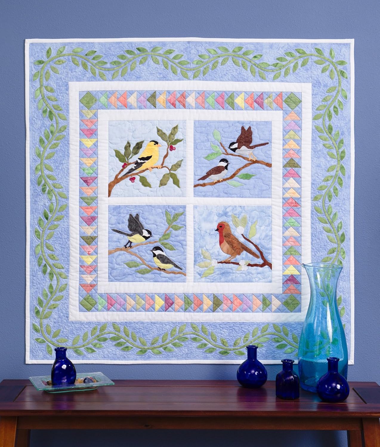 Quilt with bird designs on a wall above a wooden table with decorative items.