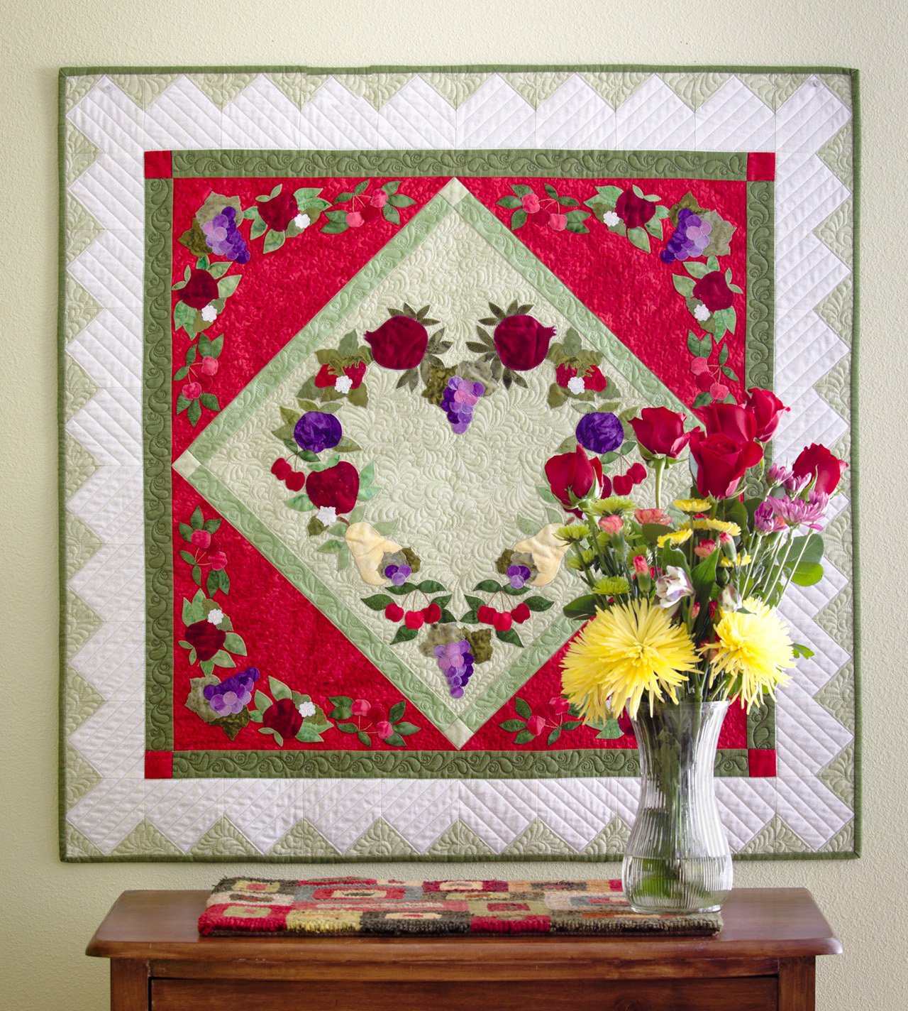Quilt with floral design on a wall above a wooden table with a vase of flowers.