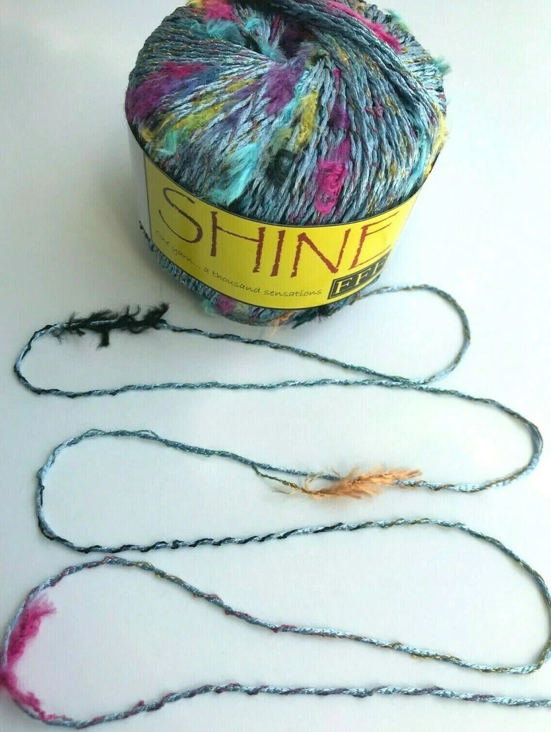 Multicolored yarn ball with 'Shine' label on a white background