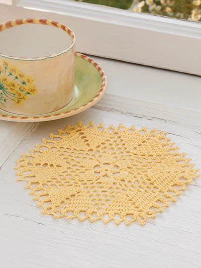 Yellow crocheted doily on a white surface with a floral teacup in the background.