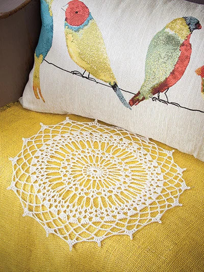 Crocheted doily on a yellow surface with embroidered bird pillows in the background