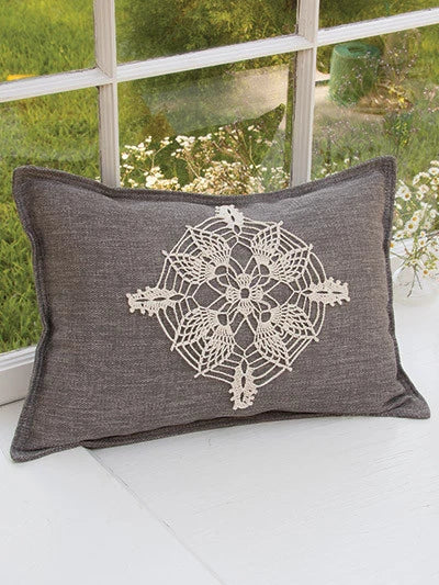 Gray pillow with a decorative white pattern on a white surface with a window in the background.