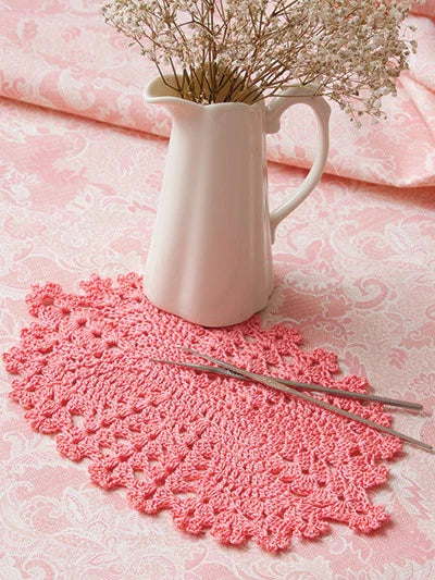 Pink crocheted doily with a white pitcher and dried flowers on a pink lace tablecloth.