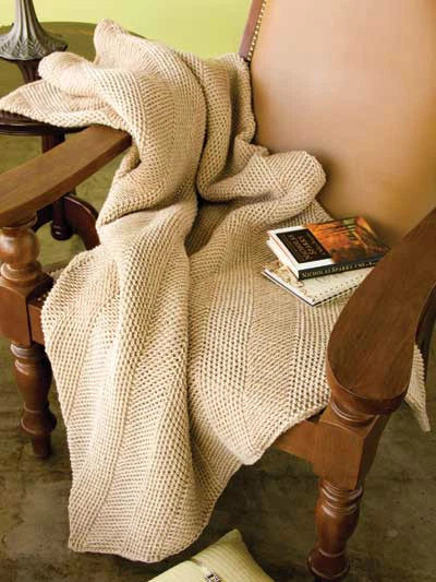Beige knitted blanket draped over a wooden chair with a book on a small table.