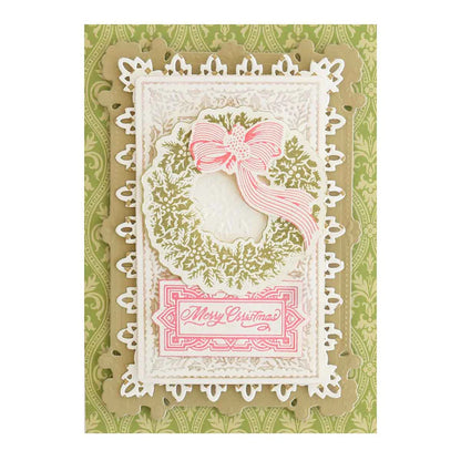 A handmade Christmas card is presented, featuring intricate die-cut designs of a wreath and floral elements. The card has a green background and white layers, accented with a pink bow and a "Merry Christmas" sentiment tag. 