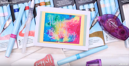 A vibrant, abstract watercolor painting of a French Bulldog's face is shown on white paper, alongside a 3-pack of "Spectrum Noir AquaTint" pens in shades of blue, purple, and pink. The pens are water-soluble and dye-based, used to create watercolor effects.