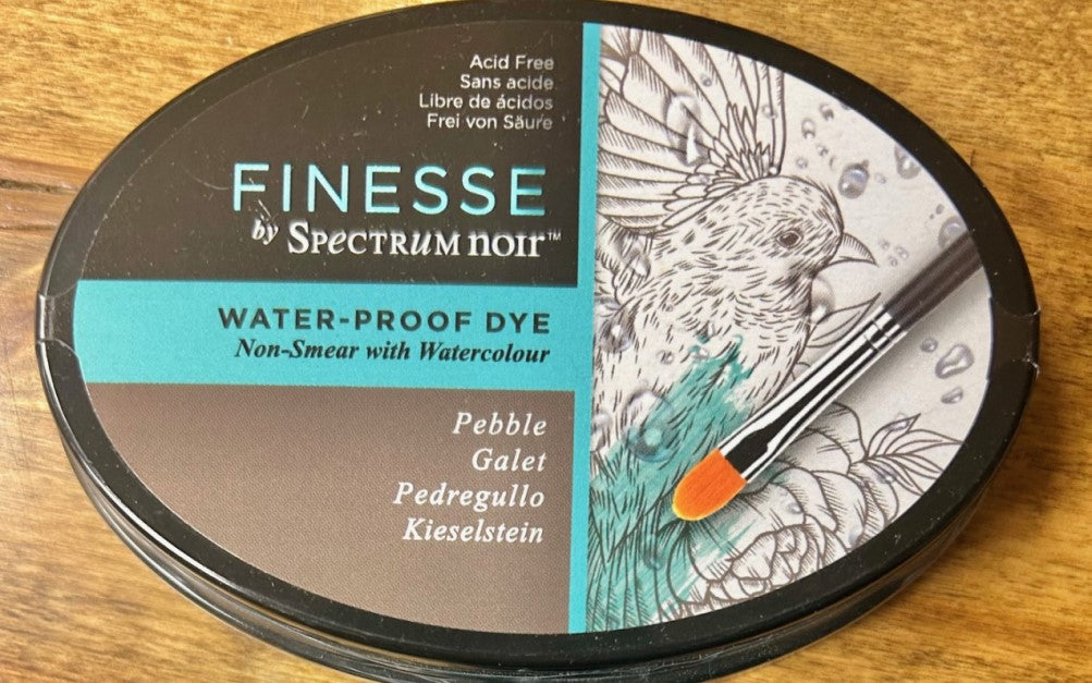 A round, dark grey Crafter's Companion Spectrum Noir Finesse water-proof dye ink pad in the color 'Pebble'. The ink pad is acid-free and designed for use with watercolour media as it is water-proof and non-smear.