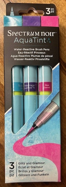 A three-pack of Spectrum Noir AquaTint water-reactive brush pens in the 'Glitz and Glamour' color set is displayed. These pens contain bright, water-soluble dye-based ink dispensed through a fine nylon brush tip. The set includes the colors Aquamarine, Amethyst, and Pink Garnet. The pens can be blended with water for various effects and are suitable for mixed media. 