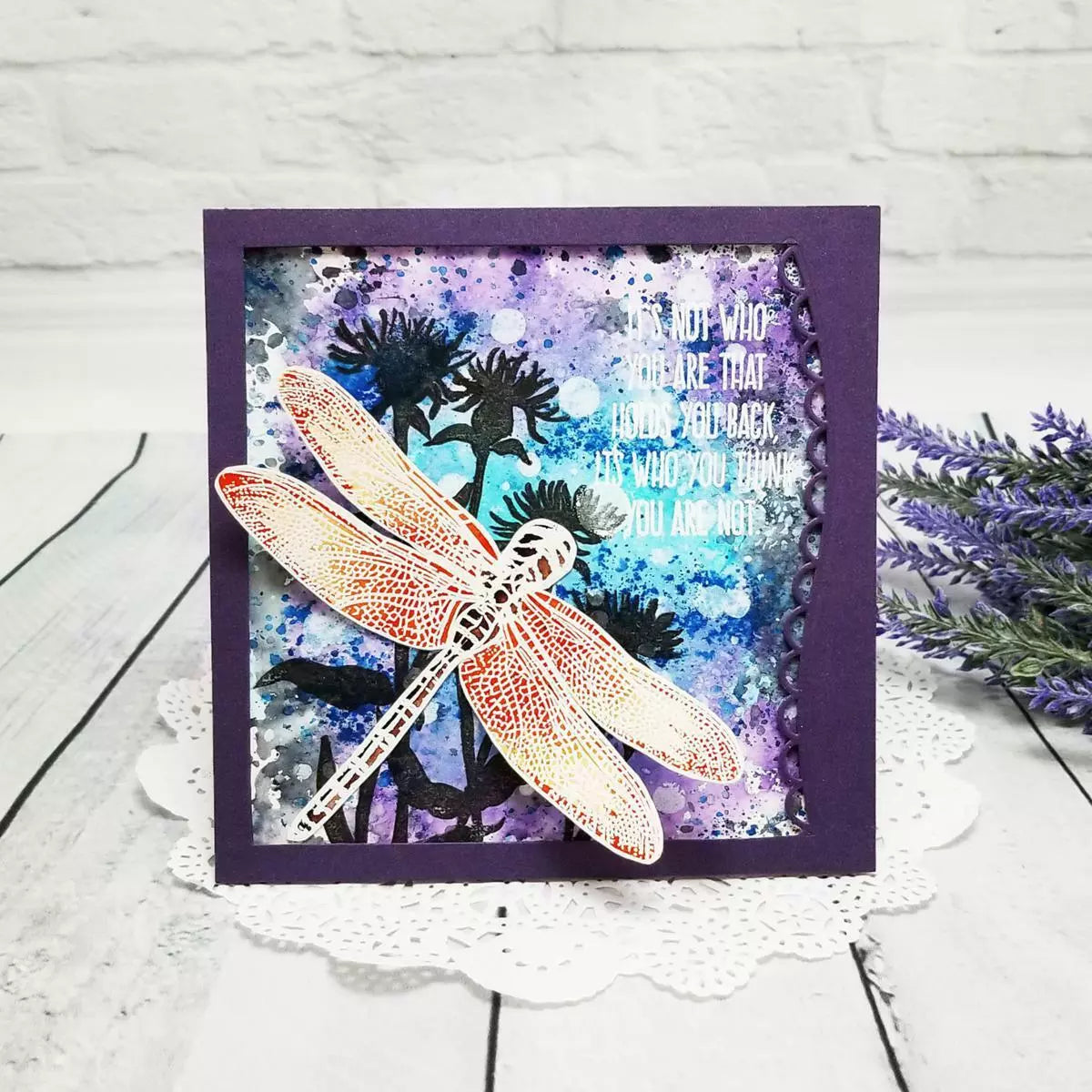 A handmade encouragement card featuring a white die-cut dragonfly with orange and yellow accents, layered over a purple and blue watercolor background. The background has silhouettes of dark flowers and a white, doily-style border on the bottom right.