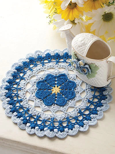 Crocheted doily with blue floral design on a table with a teacup and flowers.