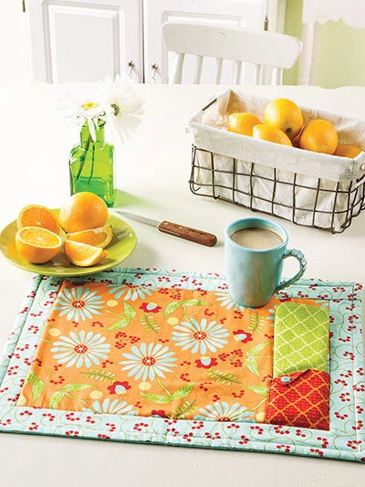 Table setting with a floral placemat, mug, plate of oranges, and basket on a white table.