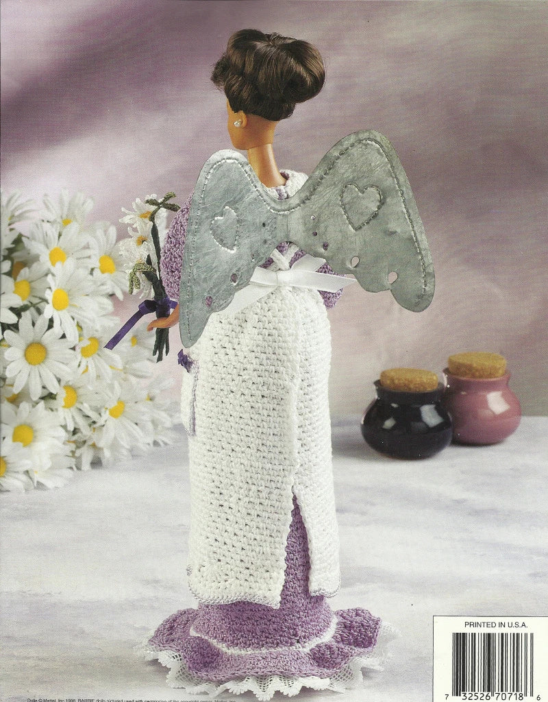 Knitted angel doll with wings and a purple dress, surrounded by flowers and jars on a textured surface.