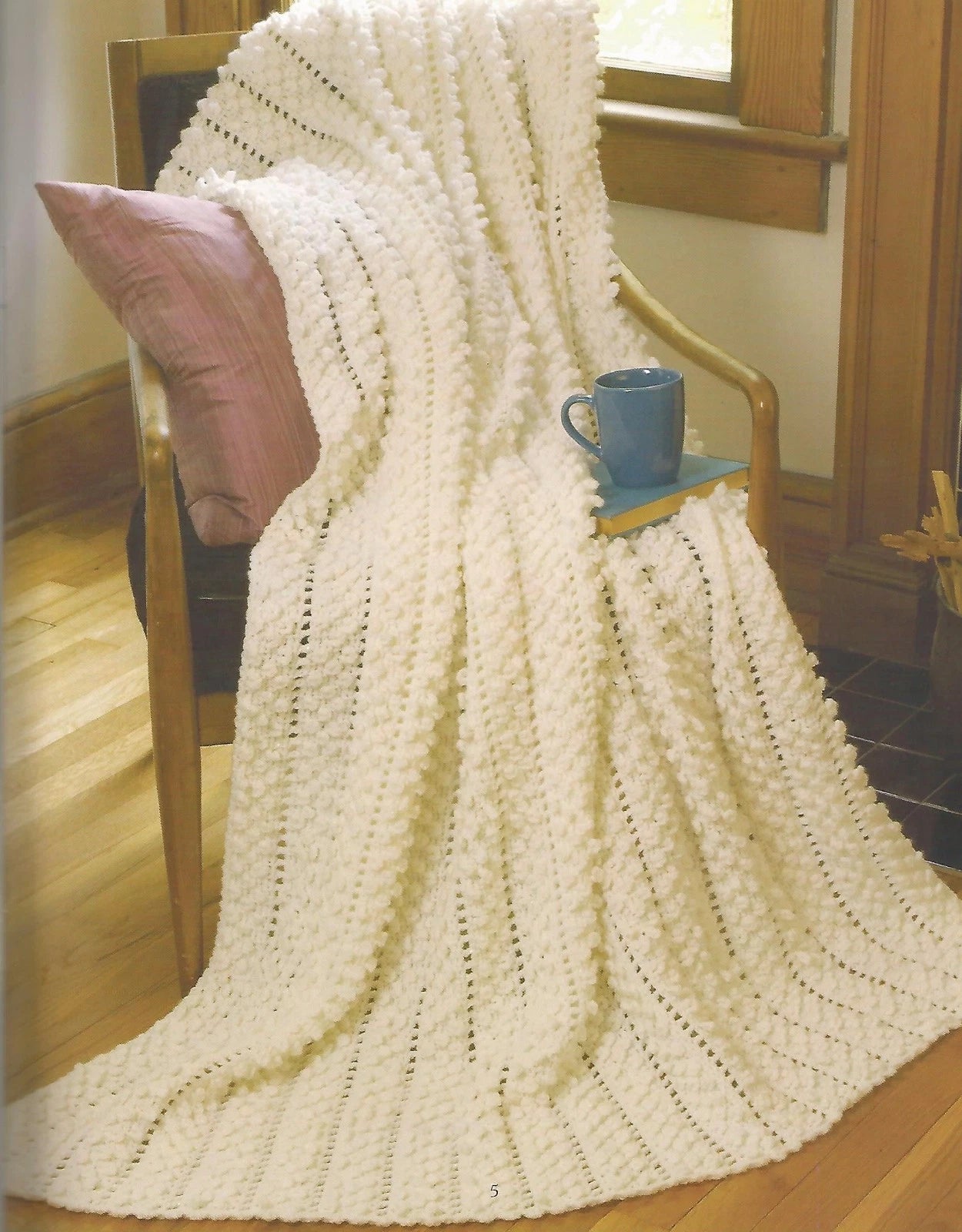 Knitted blanket draped over a chair with a cup and pillow in a cozy room.