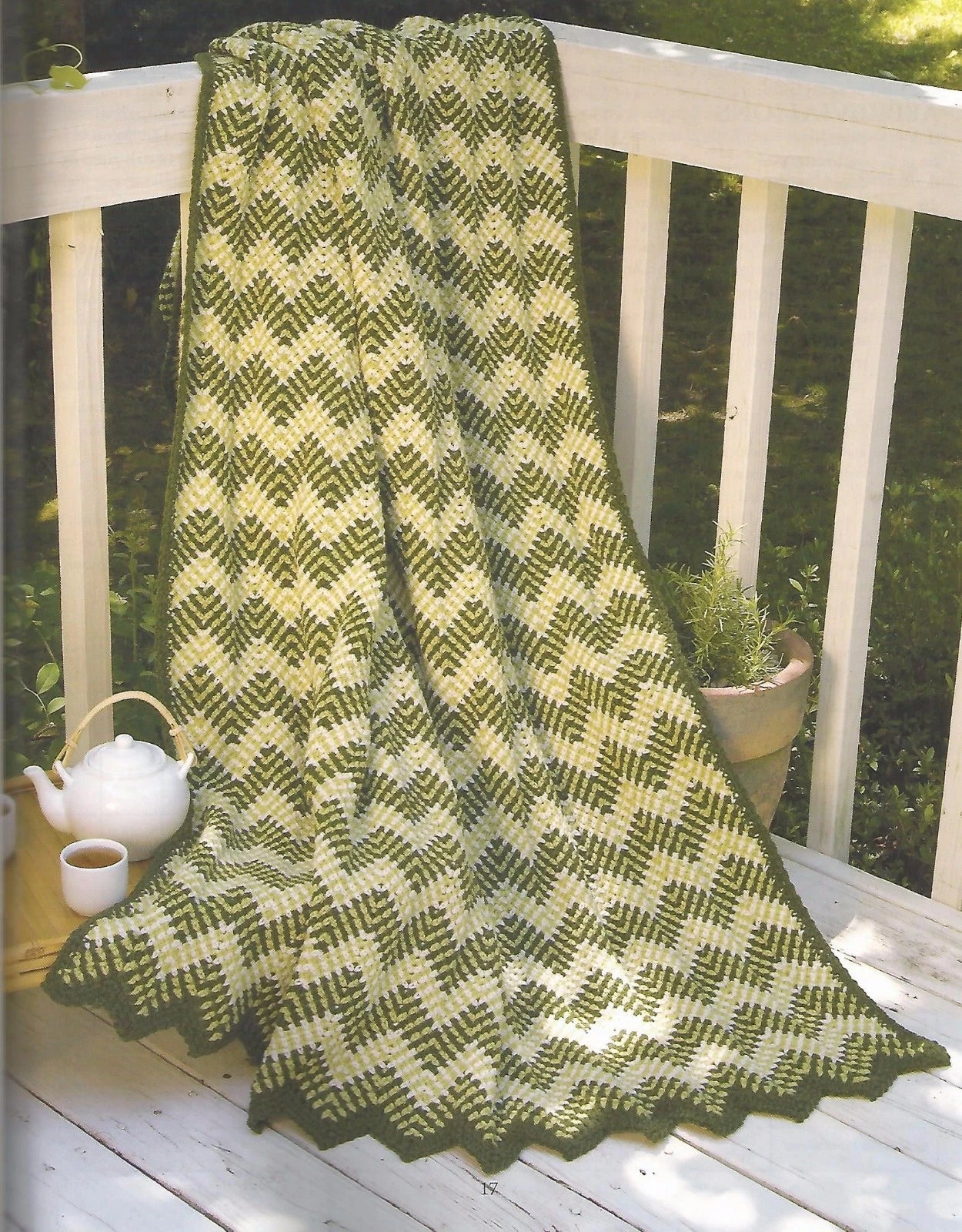 Knitted green and white blanket on a wooden railing with a teapot and cup in the background.