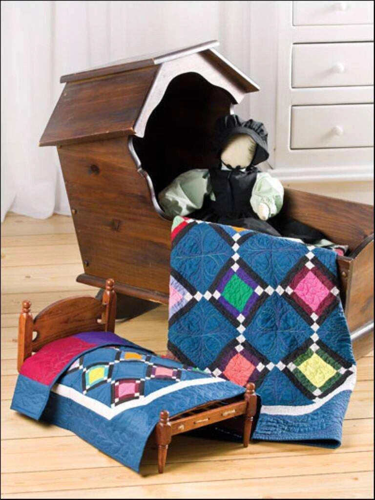 Dollhouse with miniature bed and colorful quilt, next to a wooden toy chest with dolls inside.