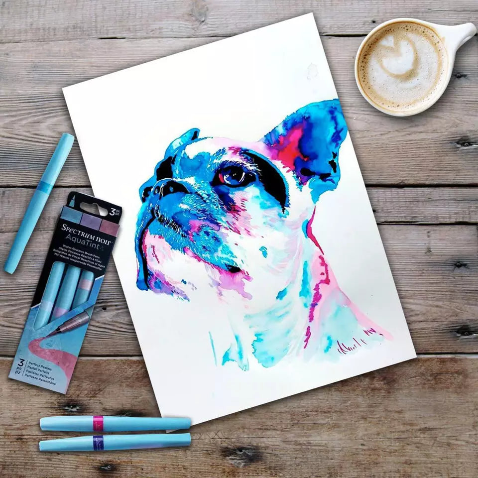 A vibrant, abstract watercolor painting of a French Bulldog's face is shown on white paper, alongside a 3-pack of "Spectrum Noir AquaTint" pens in shades of blue, purple, and pink. The pens are water-soluble and dye-based, used to create watercolor effects.