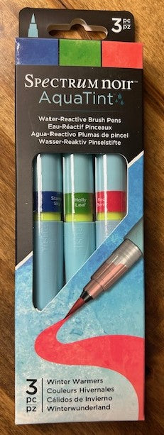 A three-pack of Spectrum Noir AquaTint water-reactive brush pens in the 'Winter Warmers' color set. The pens contain bright, water-soluble dye-based ink dispensed through a fine nylon brush tip. The set includes the colors Starry Sky, Holly Leaf, and Red Berry. These pens are suitable for mixed media and can be blended with water for various effects. 
